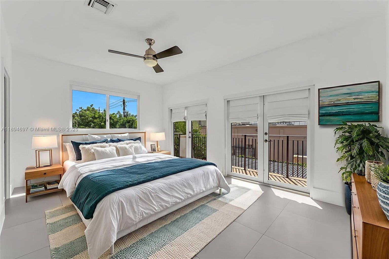 This is a bright and airy primary bedroom featuring a large bed with white linens and a teal throw blanket. The room has a modern aesthetic with white walls, tile flooring, and access to a balcony through double doors. Natural light floods the space, highlighting the clean lines and minimalist decor, including a piece of artwork and a potted plant.
