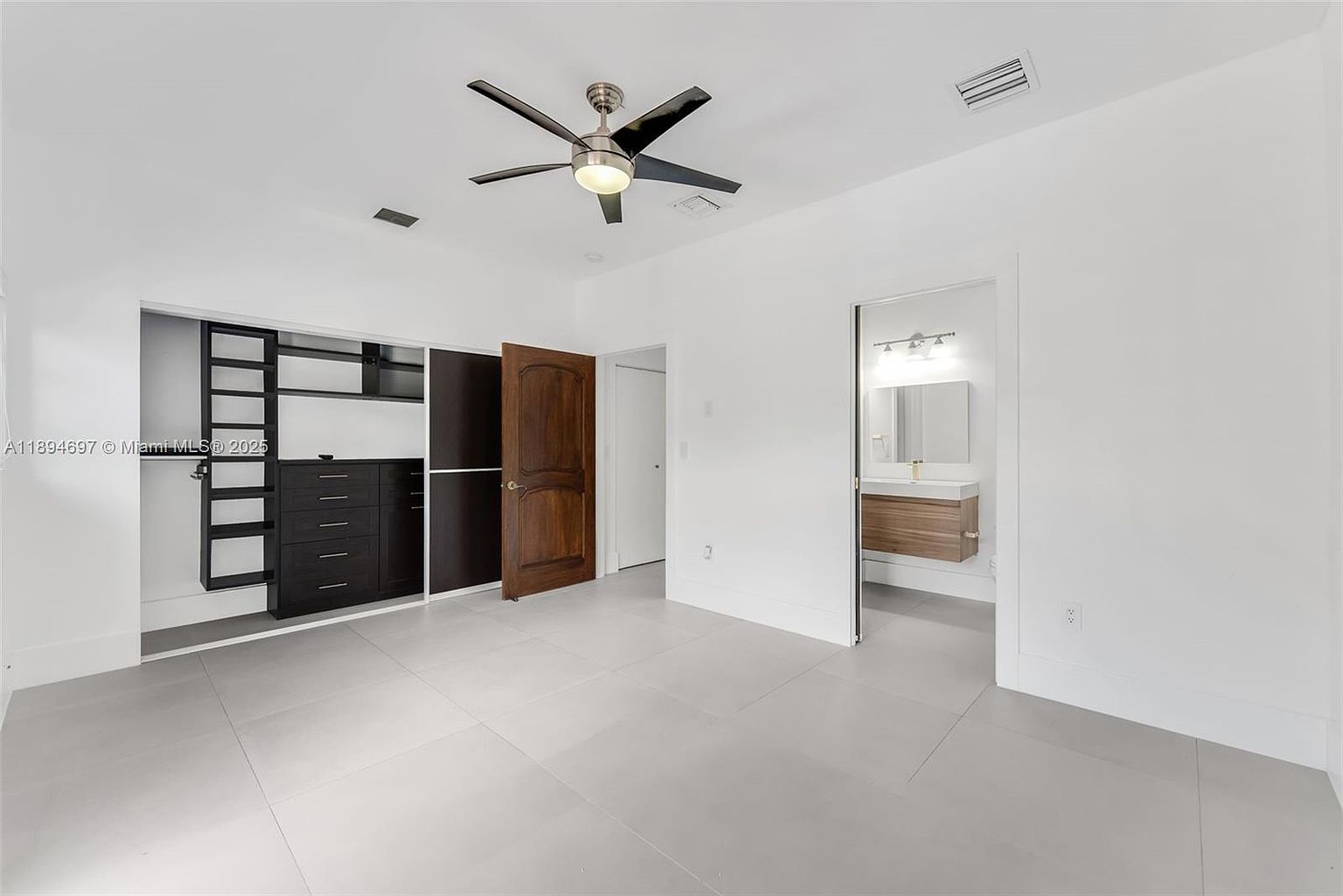 This is a bright and spacious primary bedroom featuring a modern ceiling fan and light gray tile flooring. The room includes a built-in closet system with dark wood accents and a separate walk-in closet with a dark wood door. An open doorway leads to an en-suite bathroom, enhancing the room's functionality and appeal.