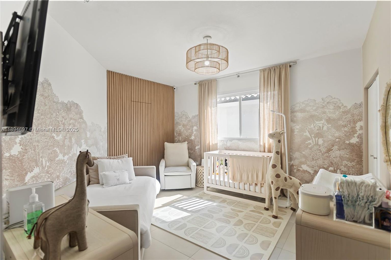 This is a cozy and well-designed nursery. The room features a crib, a comfortable daybed, and a rocking chair, creating a soothing environment. The walls are decorated with a subtle tree mural and wooden paneling, complemented by a stylish light fixture and a patterned rug, enhancing the room's serene and inviting atmosphere.