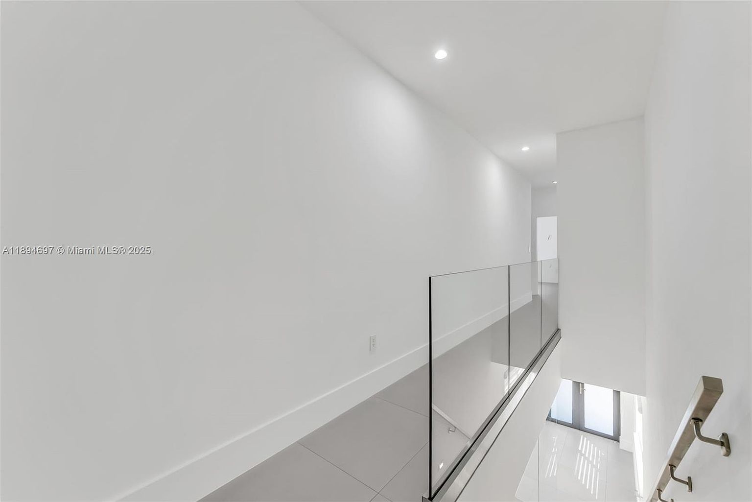 This is an interior shot of a modern hallway and staircase. The hallway features sleek, light gray tile flooring and a glass railing, creating an open and airy feel. The walls are painted a clean white, enhancing the brightness of the space, and recessed lighting is visible in the ceiling.