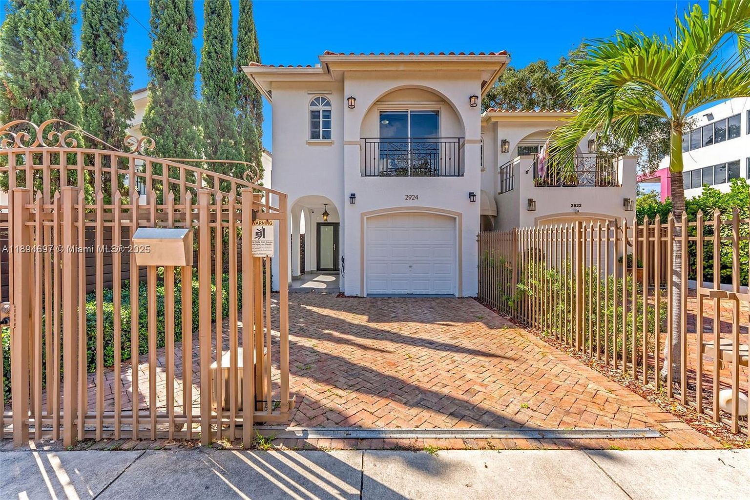 This is a front view of a two-story Mediterranean-style townhouse. The house features a white facade, a red tile roof, and an arched balcony above the garage. A brick driveway leads to the garage, and a decorative metal gate encloses the property, adding to its curb appeal.