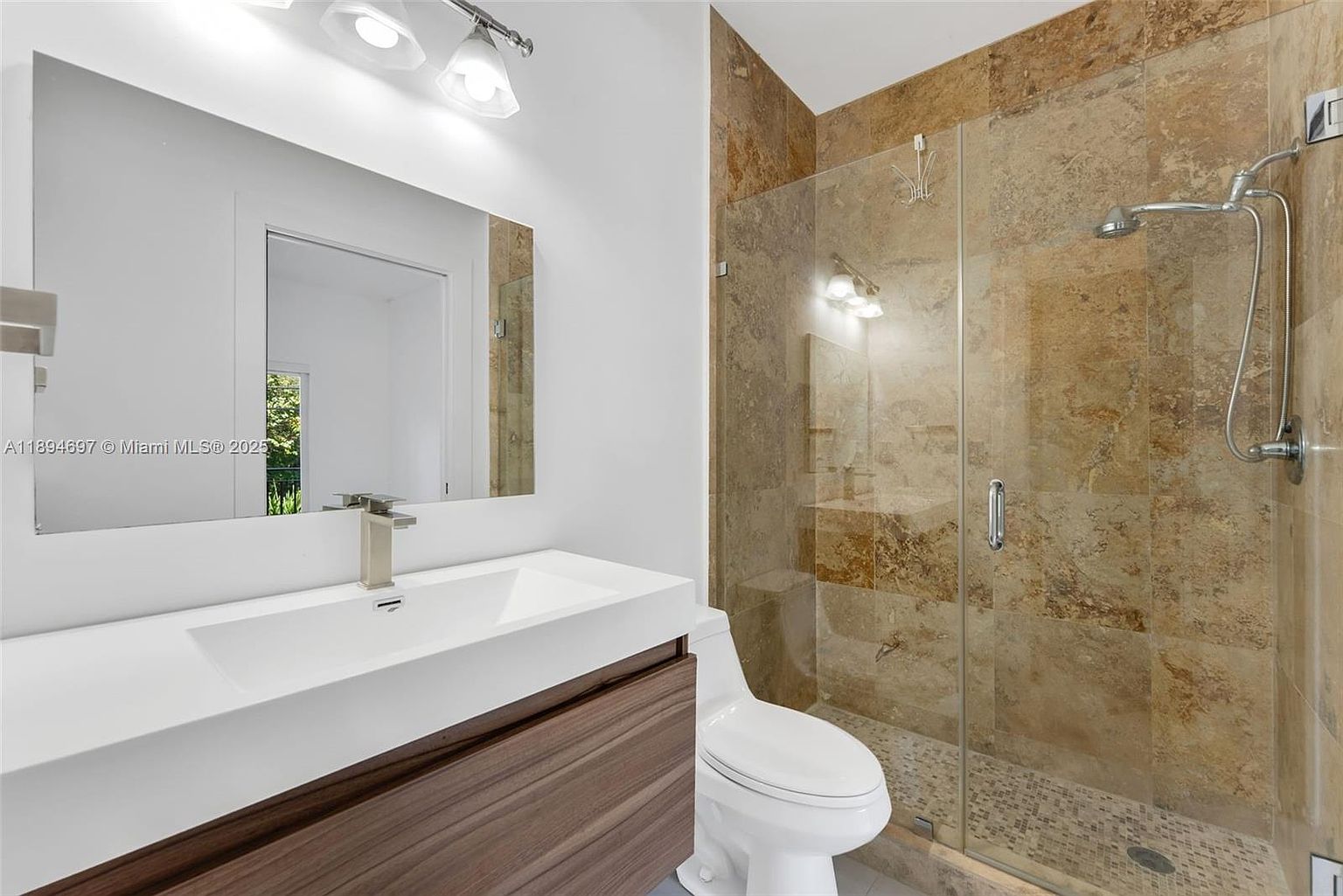 This is a well-lit primary bathroom featuring a modern vanity with a white countertop and dark wood cabinetry. A large mirror hangs above the sink, reflecting the natural light. The shower is enclosed with glass and features travertine tile and a pebble floor, creating a spa-like atmosphere.