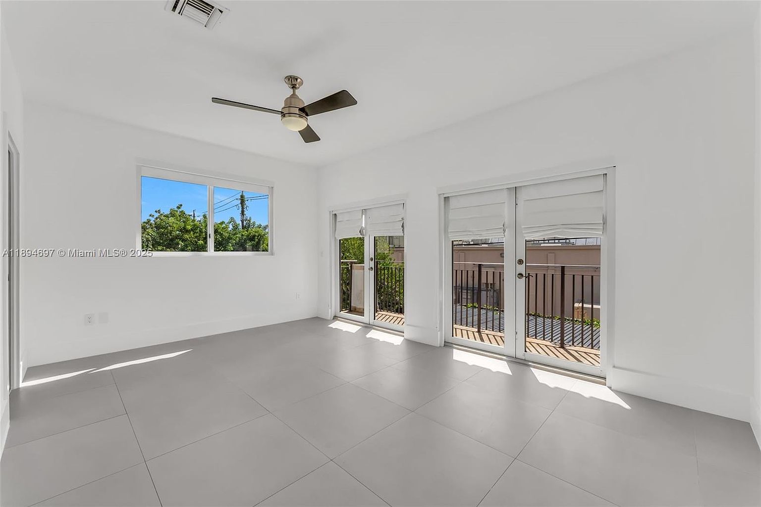 This is a bright and airy primary bedroom featuring a ceiling fan, a large window with a view of greenery, and double doors leading to a balcony. The room has a minimalist aesthetic with white walls and light gray tile flooring, creating a clean and spacious feel. The perspective is from the corner of the room, showcasing the window and balcony access.