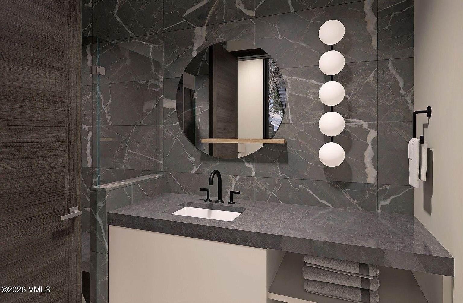 This modern bathroom features a sleek vanity with a dark stone countertop and a white undermount sink. The wall is finished with large-format, dark veined stone tiles, complemented by a circular mirror and a vertical light fixture with five spherical bulbs. A glass-enclosed shower area is visible to the left, and a towel rack with a white towel is mounted on the right wall.