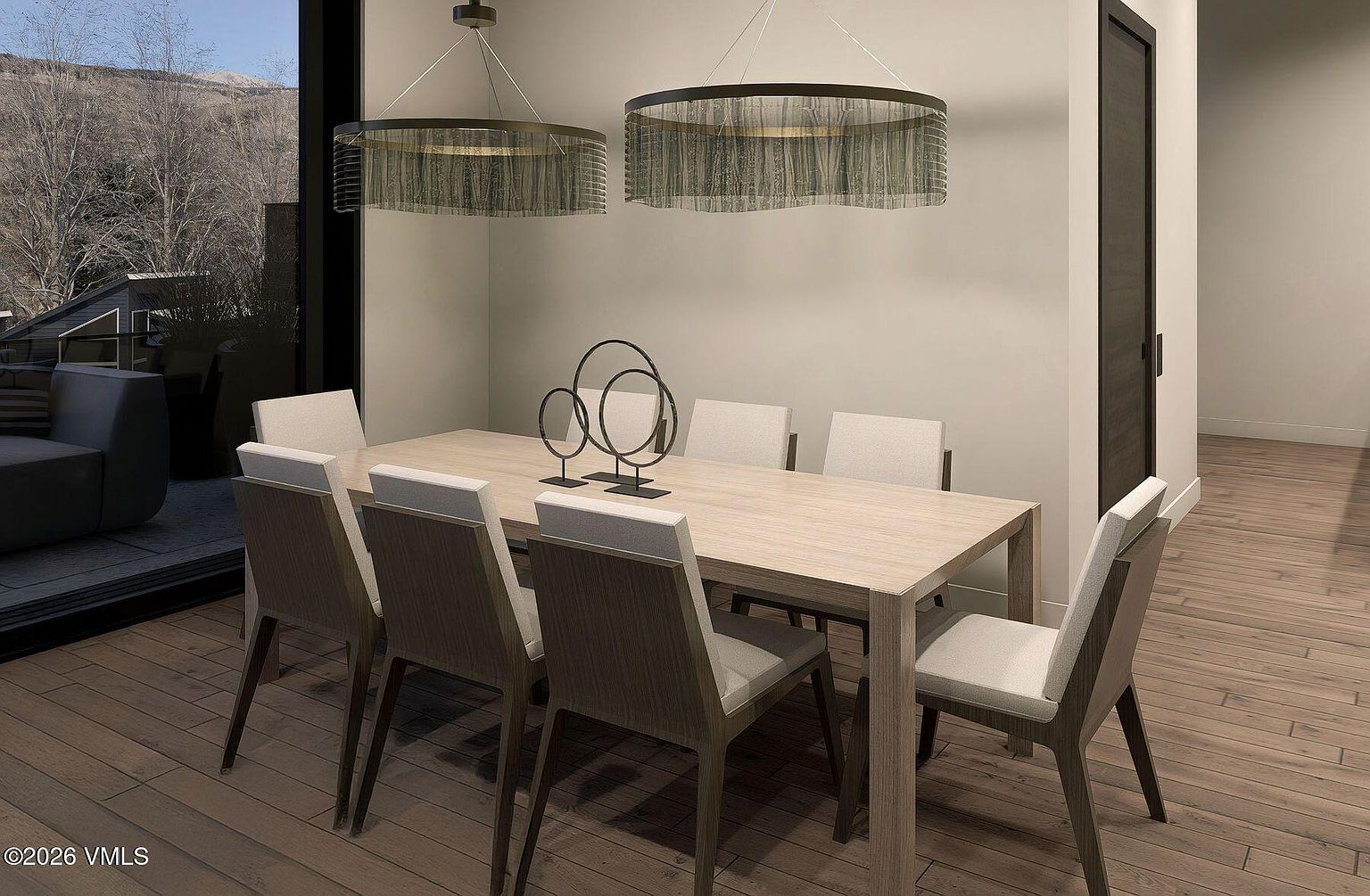 This contemporary dining room features a light wood table surrounded by six upholstered chairs, centered beneath two large, circular drum-style pendant lights. To the left, expansive glass panels provide a view of an outdoor deck and mountain scenery, while the room's neutral walls and wood flooring create a cohesive, modern aesthetic. A sculptural metal centerpiece sits on the table, adding a refined artistic touch to the minimalist space.
