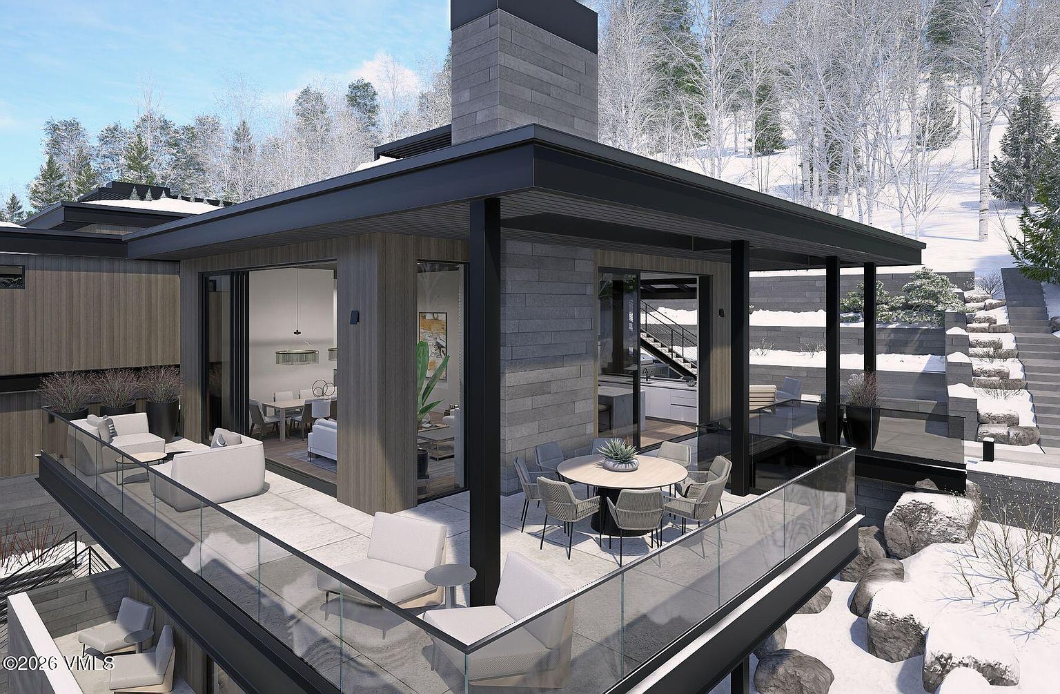 This modern mountain residence features an expansive outdoor deck designed for seamless indoor-outdoor living, complete with a dining area and lounge seating. The structure showcases a contemporary aesthetic with a prominent stone-clad chimney breast, dark metal framing, and wood-paneled exterior walls. Large glass sliding doors provide a clear view into the interior living space, while the surrounding snowy landscape and tiered stone retaining walls create a serene, high-end alpine atmosphere.