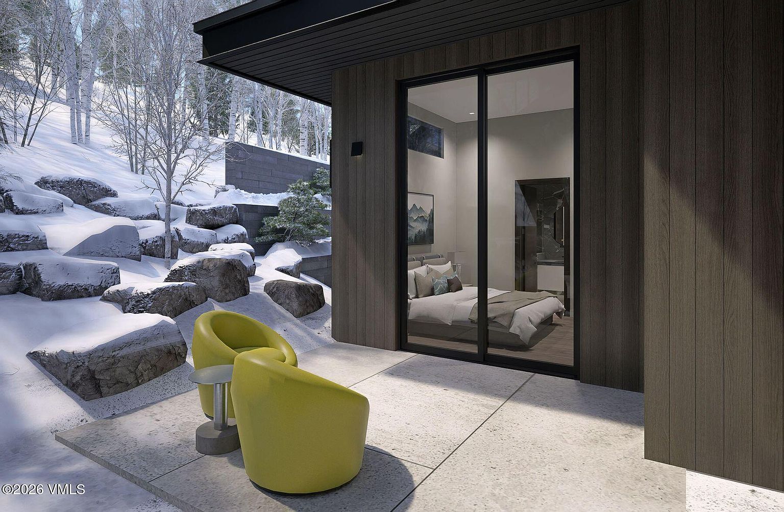 This modern patio space features two vibrant yellow chairs and a small side table, set against a backdrop of a snowy, rocky landscape. Large glass sliding doors provide a seamless transition into a bedroom, highlighting the contemporary architectural design and the integration of indoor and outdoor living. The dark wood-paneled exterior walls contrast sharply with the bright, wintry environment, creating a sophisticated and serene atmosphere.