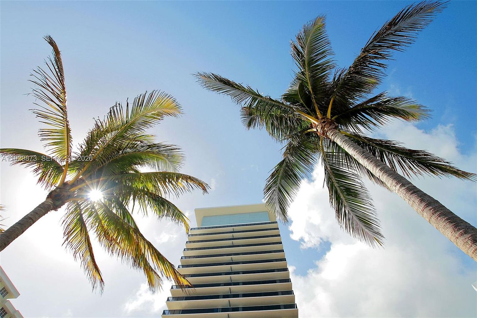 The image showcases a modern high-rise building framed by two palm trees against a bright blue sky with scattered clouds. The building features a sleek design with multiple balconies, creating a layered effect. The perspective is from a low angle, emphasizing the height of the building and the tropical setting, suggesting a luxurious and contemporary living environment.
