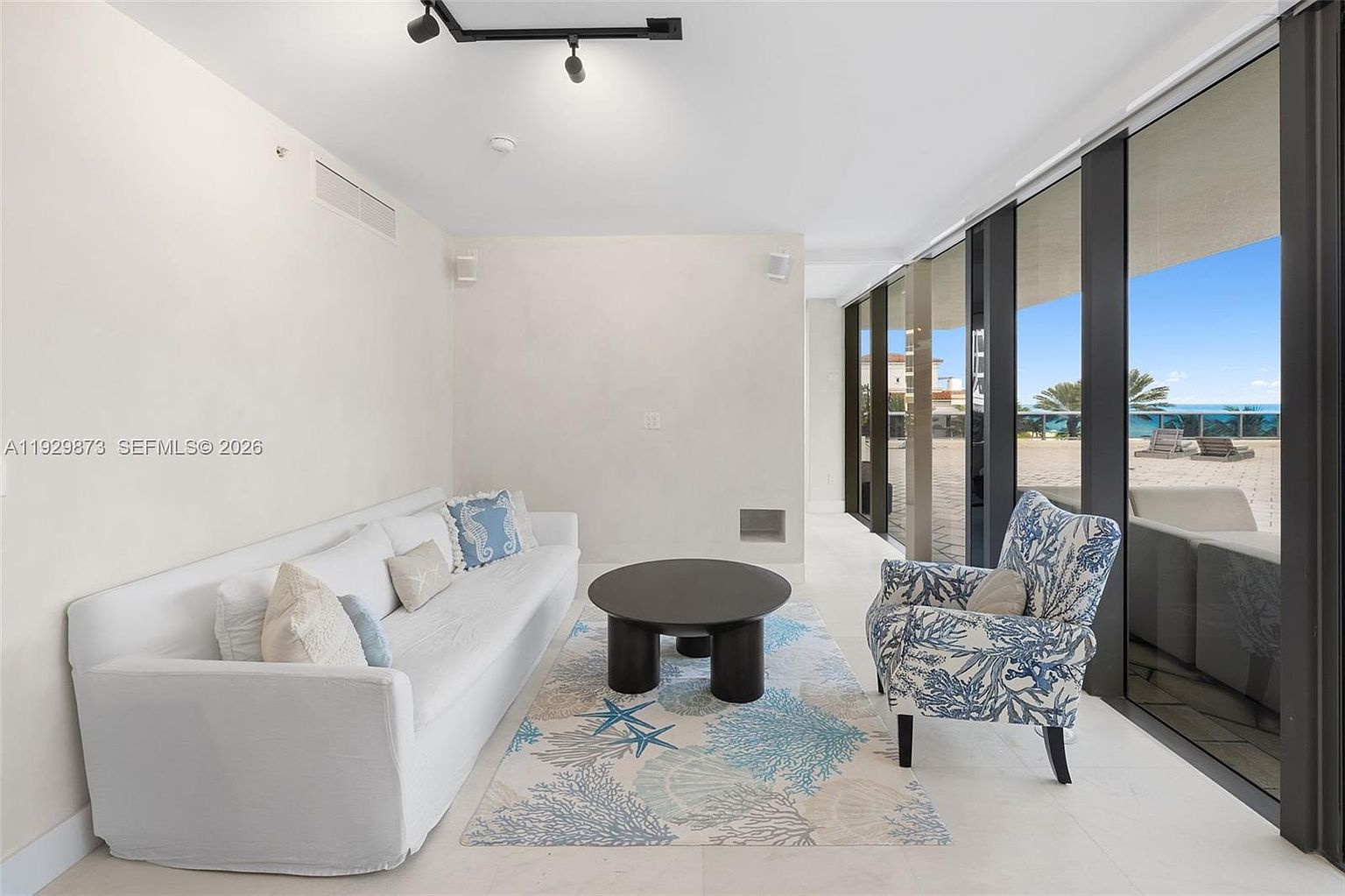 This is an interior shot of a living room featuring a white sofa with decorative pillows, a patterned armchair, and a round coffee table on a patterned rug. Large sliding glass doors offer a view of the ocean and a patio area, creating a bright and airy atmosphere. The room is decorated in a coastal style with a neutral color palette.