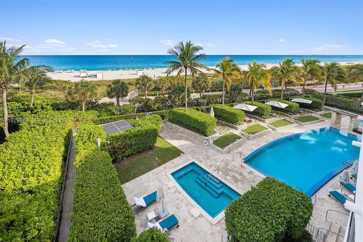 This aerial view showcases a luxurious pool and spa area adjacent to a pristine beach. Lush green landscaping surrounds the pool, providing privacy and a tropical feel. The turquoise water of the pool contrasts beautifully with the white sand and blue ocean in the background, creating an inviting and serene atmosphere.