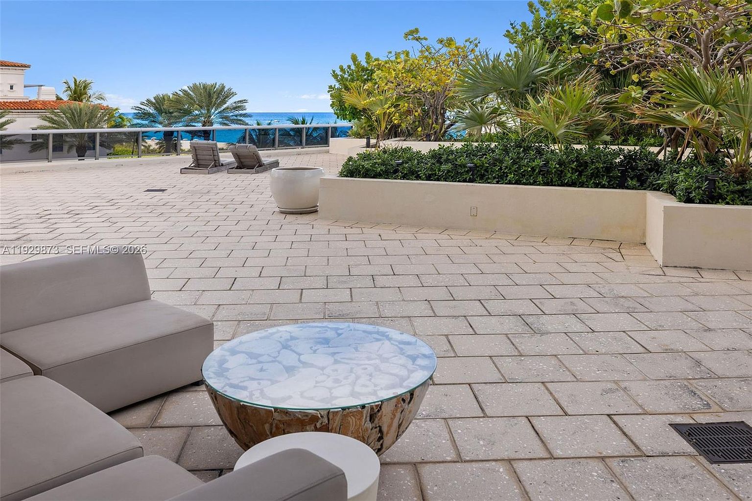 This image showcases a luxurious outdoor patio or balcony area, featuring comfortable lounge seating arranged around a unique, round coffee table with a glass top and a natural wood base. The space is paved with light-colored stone tiles and is bordered by lush greenery and a low wall, offering a partial view of the ocean and sky in the background. The overall impression is one of relaxation and upscale outdoor living.