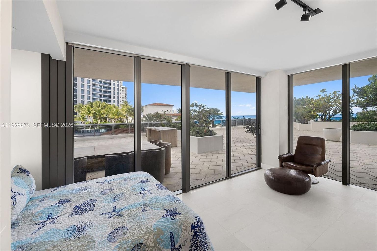 This primary bedroom features a large sliding glass door that opens onto a spacious balcony with outdoor seating and ocean views. The room is decorated with a bed featuring a blue and white patterned quilt, a leather chair with an ottoman, and white tile flooring. The overall impression is one of luxury and relaxation, with a focus on the outdoor living space and natural light.