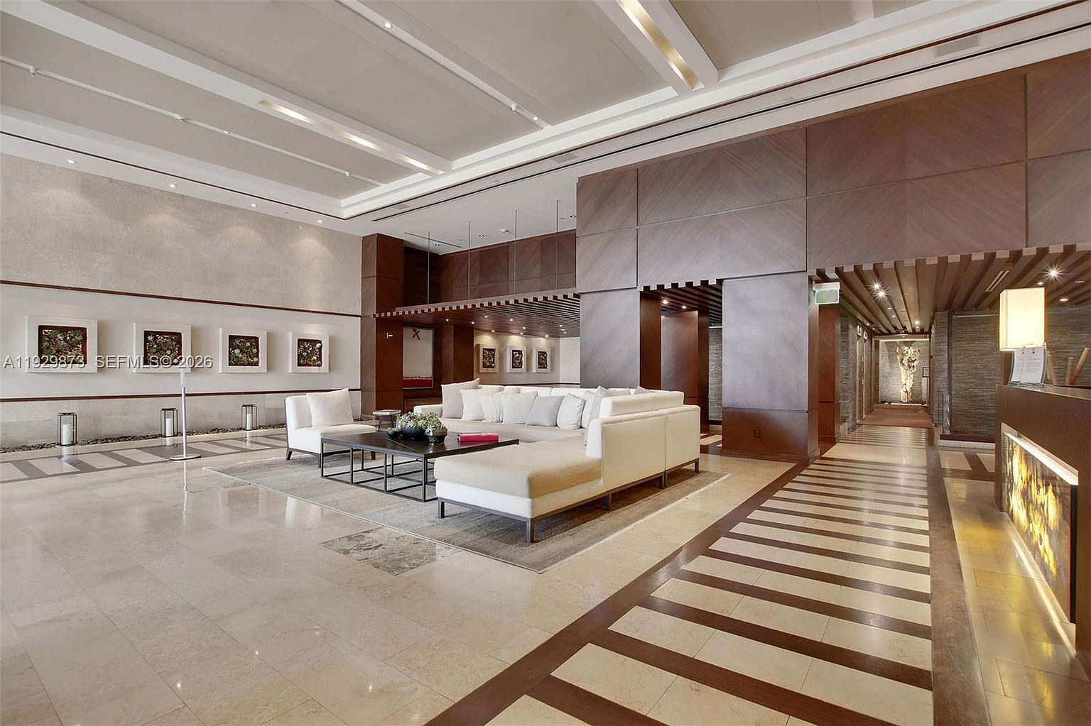 This is a spacious and luxurious living room area, likely in a high-end residential building or hotel. The room features a large, modern sectional sofa, a sleek coffee table, and stylish artwork on the walls. The flooring is a combination of light-colored tile and dark wood accents, creating a sophisticated and inviting atmosphere.