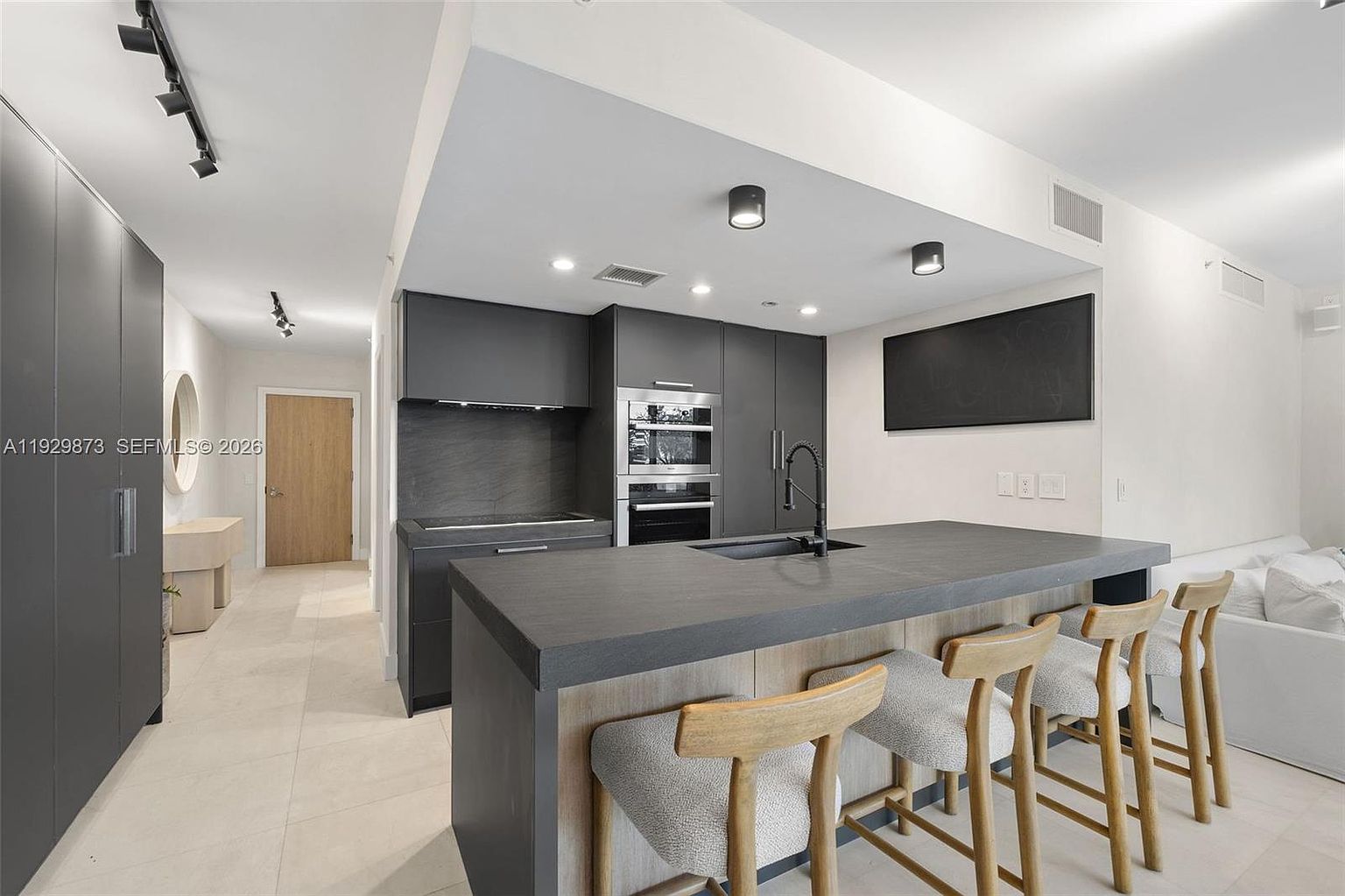 This is a modern kitchen featuring sleek, dark gray cabinetry and a contrasting light wood island with a dark countertop. Stainless steel appliances are integrated into the cabinetry, and the island is complemented by three bar stools with light-colored upholstery. The space is well-lit with recessed lighting and track lighting, creating a clean and contemporary atmosphere.