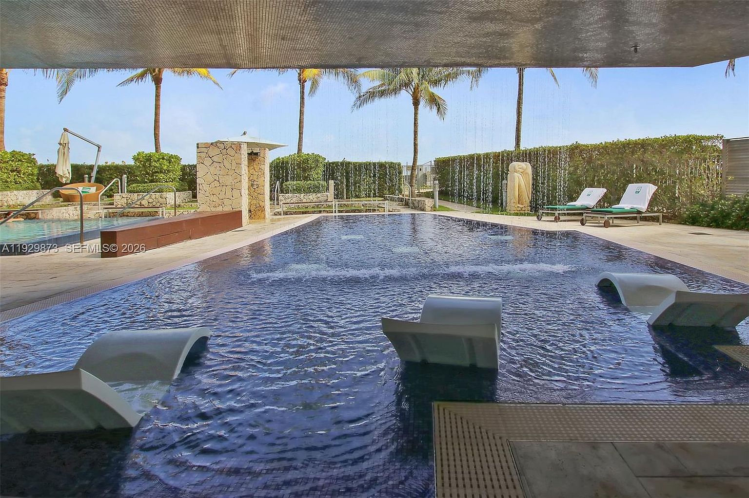 This image showcases a luxurious pool area, featuring a dark-tiled pool with built-in lounge chairs partially submerged in the water. The pool is surrounded by a well-maintained deck, lush greenery, and palm trees, creating a serene and upscale atmosphere. Water features add to the ambiance, suggesting a high-end resort-style living experience.