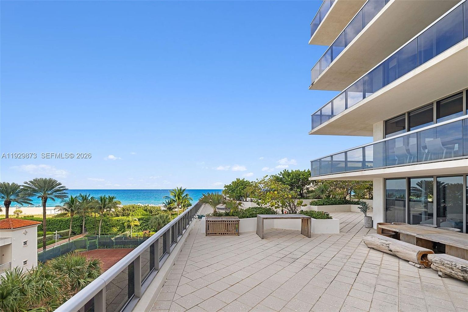 This image showcases a luxurious balcony or patio area with a stunning ocean view. The space features modern design elements, including glass railings, neutral-toned paving stones, and contemporary outdoor furniture. Lush greenery and palm trees add to the tropical ambiance, creating an inviting and upscale outdoor living space.