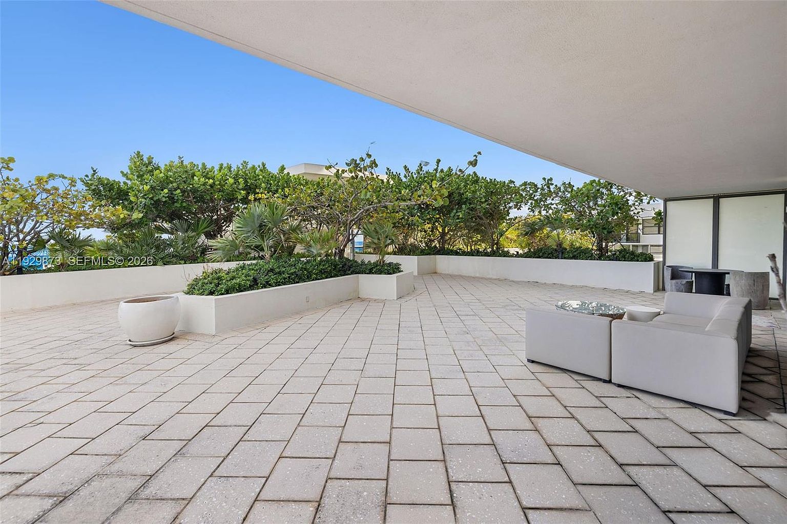 This image showcases a spacious outdoor patio or balcony area. The flooring is composed of light-colored paving stones, and the perimeter is lined with raised planters filled with greenery, offering privacy and a touch of nature. A modern outdoor seating arrangement is visible, suggesting a comfortable space for relaxation and entertaining.