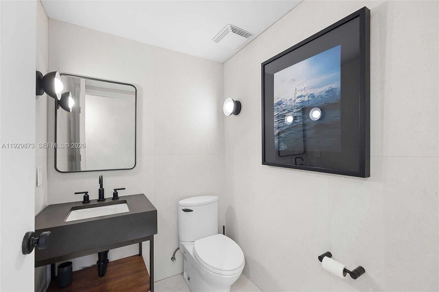 This is a modern guest bathroom featuring a sleek, dark-colored vanity with a white sink and black fixtures. A minimalist mirror hangs above the vanity, complemented by modern sconce lighting. The toilet is white and the walls are tiled in a light neutral tone, with a framed art piece adding a touch of sophistication.