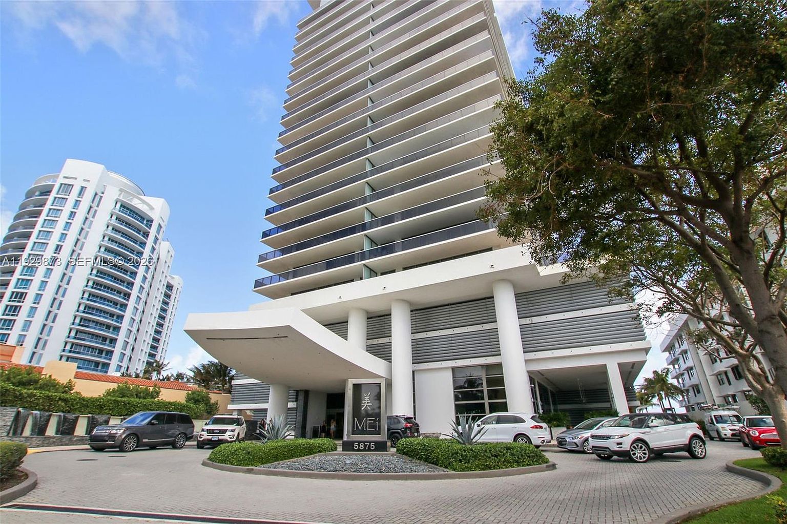 The image showcases the grand entryway of a modern high-rise building. A circular driveway leads to the entrance, which is marked by a sleek sign and landscaping. The building features a contemporary design with multiple balconies and a prominent porte-cochère, creating an impressive and welcoming arrival experience.