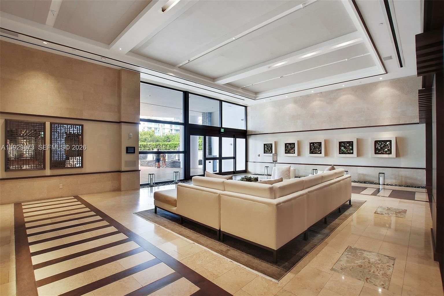This is a well-lit interior shot of a modern living room or lobby area. The space features a large, light-colored sectional sofa, marble flooring with dark wood accents, and contemporary artwork on the walls. Large windows provide natural light and a view to the outside, creating an inviting and sophisticated atmosphere.