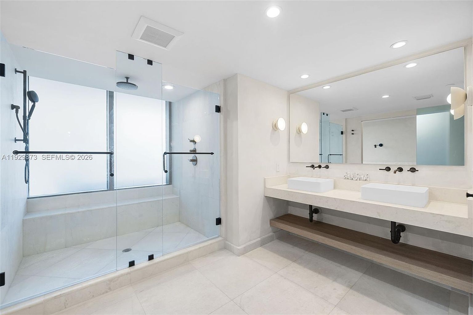 This is a bright and modern primary bathroom featuring a glass-enclosed shower with black hardware and a double vanity with vessel sinks. The space is illuminated by recessed lighting and sconces, creating a clean and luxurious atmosphere. The perspective is from the doorway, showcasing the entire bathroom layout.