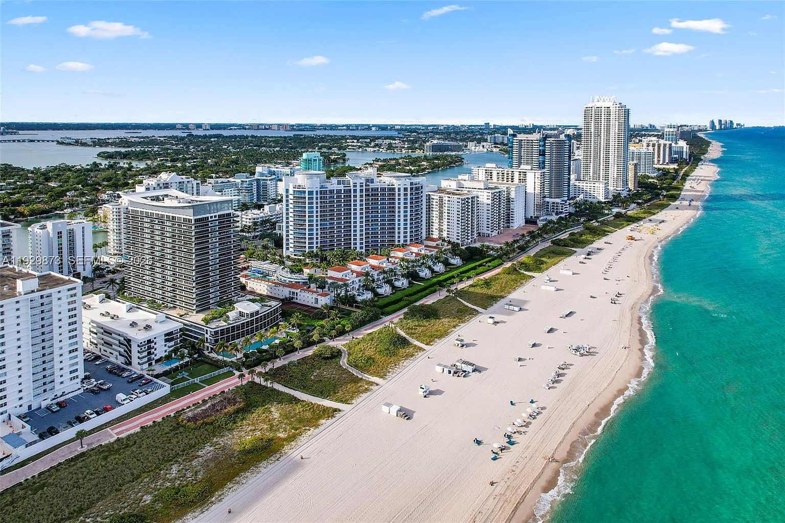 This aerial view showcases a stunning coastal property with a pristine sandy beach meeting turquoise ocean waters. Modern high-rise buildings line the coast, offering luxurious living with ocean views. Lush greenery separates the buildings from the beach, creating a serene and desirable environment.
