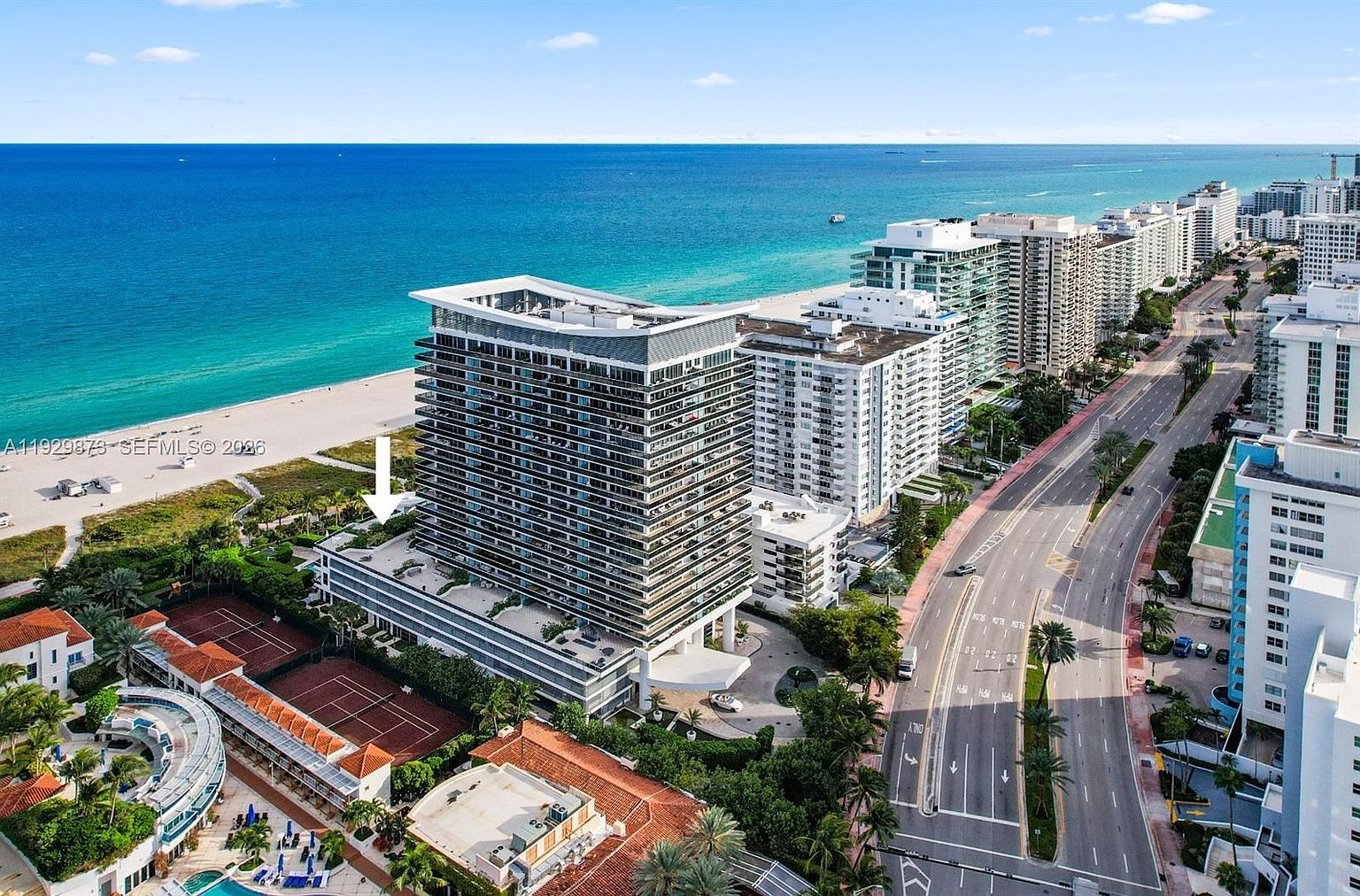 This aerial view showcases a luxury beachfront property with a modern high-rise condominium building as the focal point. The building features multiple balconies and is surrounded by lush landscaping, including tennis courts and a pool area. The turquoise ocean and a wide sandy beach provide a stunning backdrop, complemented by a well-maintained road lined with palm trees.