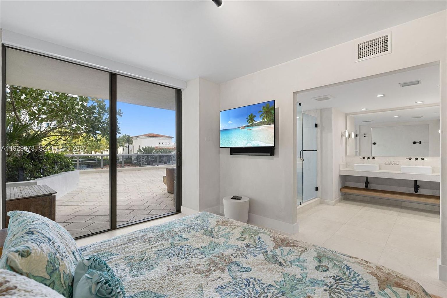 This is a primary bedroom featuring a sliding glass door leading to a patio, a wall-mounted television, and an ensuite bathroom. The bedroom is decorated in a modern style with a light color palette, creating a bright and airy atmosphere. The bed has a patterned quilt and decorative pillows.