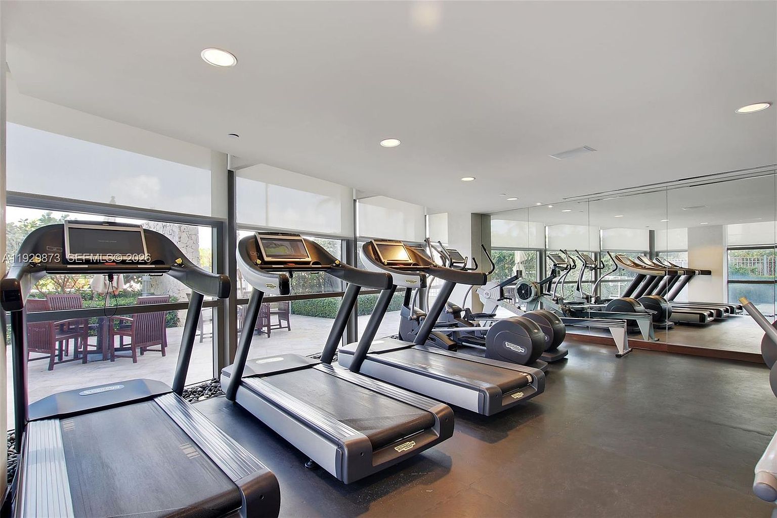 This is a well-equipped home gym featuring multiple treadmills, elliptical machines, and a rowing machine. Large windows provide natural light and a view of an outdoor patio area. The flooring is a dark, durable material, and mirrors line one wall to enhance the sense of space and provide visual feedback during workouts.