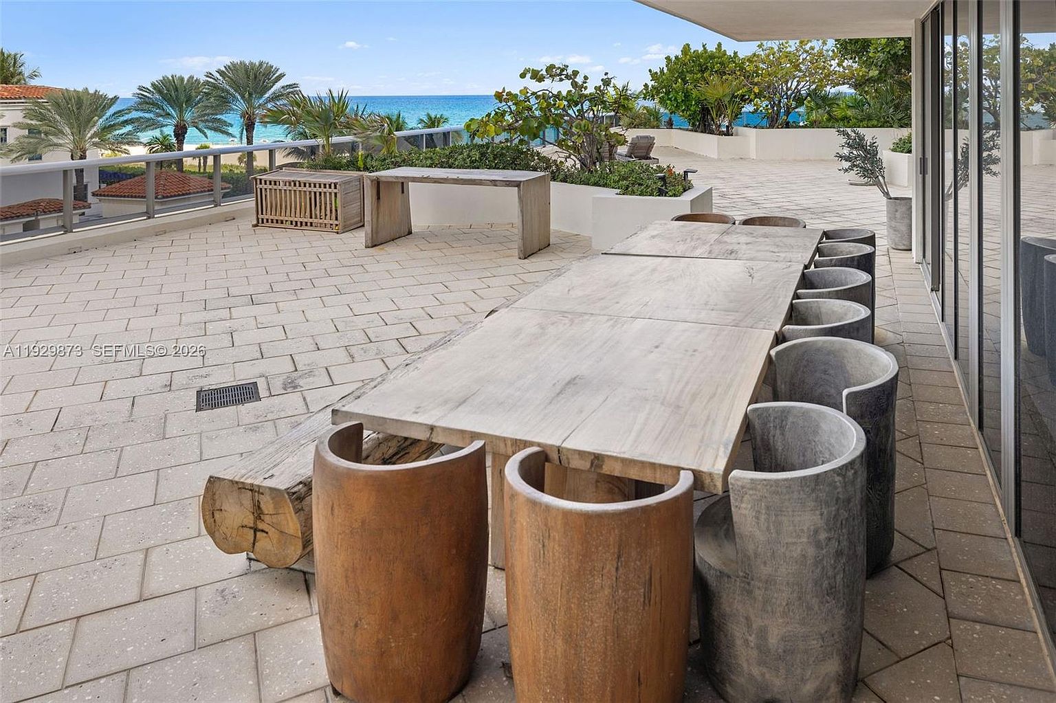 This image showcases a spacious outdoor patio or balcony area, featuring a large wooden dining table with unique log-style chairs. The patio is paved with light-colored stone tiles, and the area offers a view of the ocean and palm trees in the distance. The setting suggests a luxurious and relaxing outdoor living space.