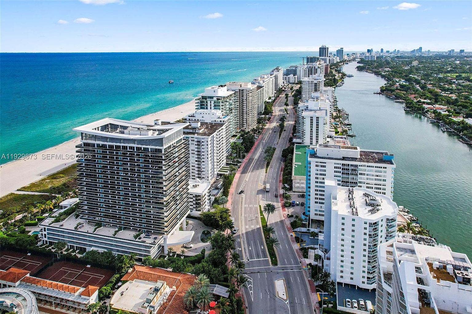This aerial view showcases a stunning coastal cityscape, featuring a line of high-rise buildings along a pristine beach and a parallel waterway. The turquoise ocean contrasts beautifully with the white sandy beach and the lush greenery lining the waterway, creating an inviting and luxurious atmosphere. The image captures the essence of upscale coastal living.
