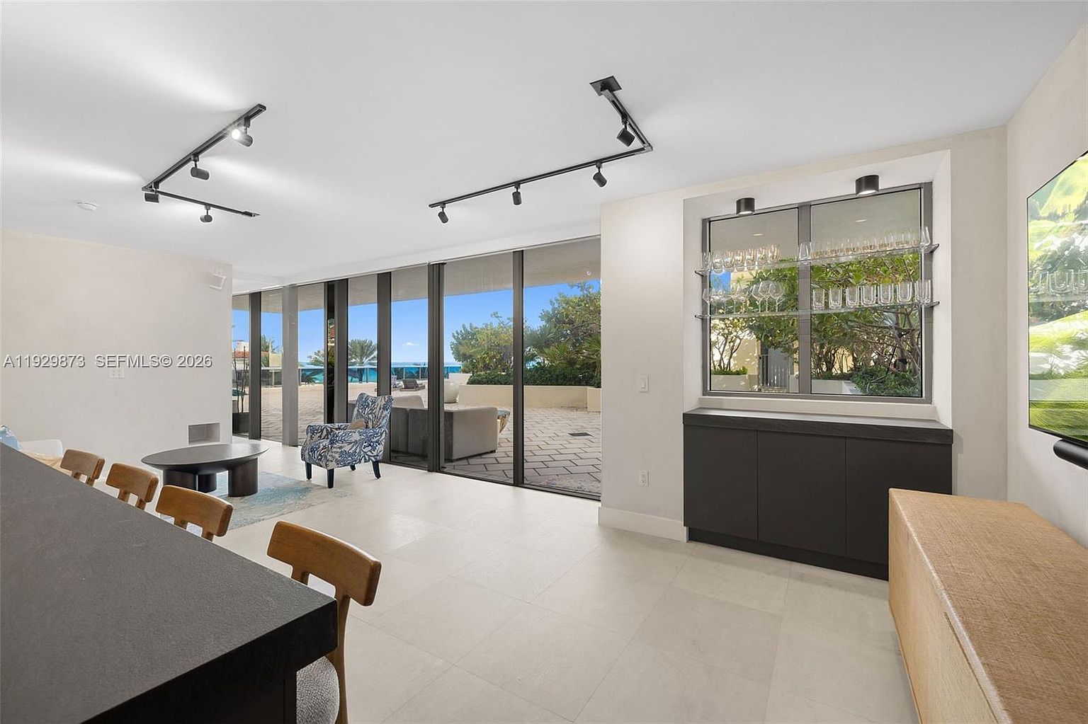 This is an interior shot of a modern living room featuring a sleek design with a view of the ocean. The room includes a bar area with glass storage, contemporary furniture, and large sliding glass doors that open to a patio. The color palette is neutral, creating a bright and airy atmosphere.