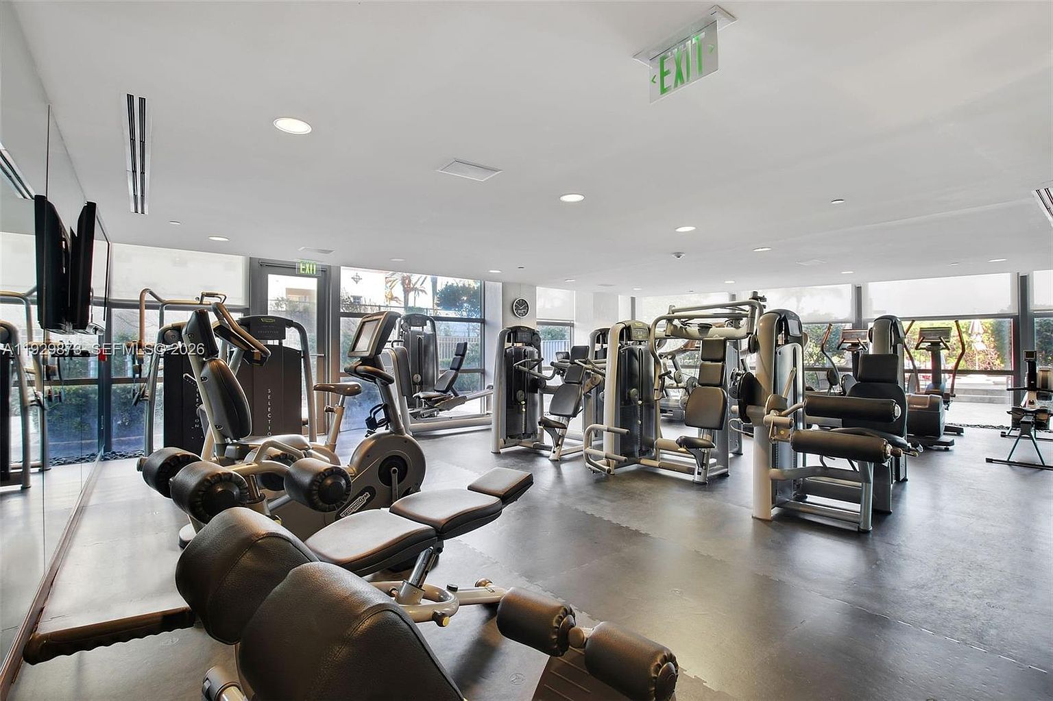 This is a well-equipped home gym featuring various exercise machines, including weight machines, stationary bikes, and treadmills. The room has large windows providing natural light and a view of the outdoors. The flooring is a dark, durable material, and the overall impression is a clean and functional space for fitness.