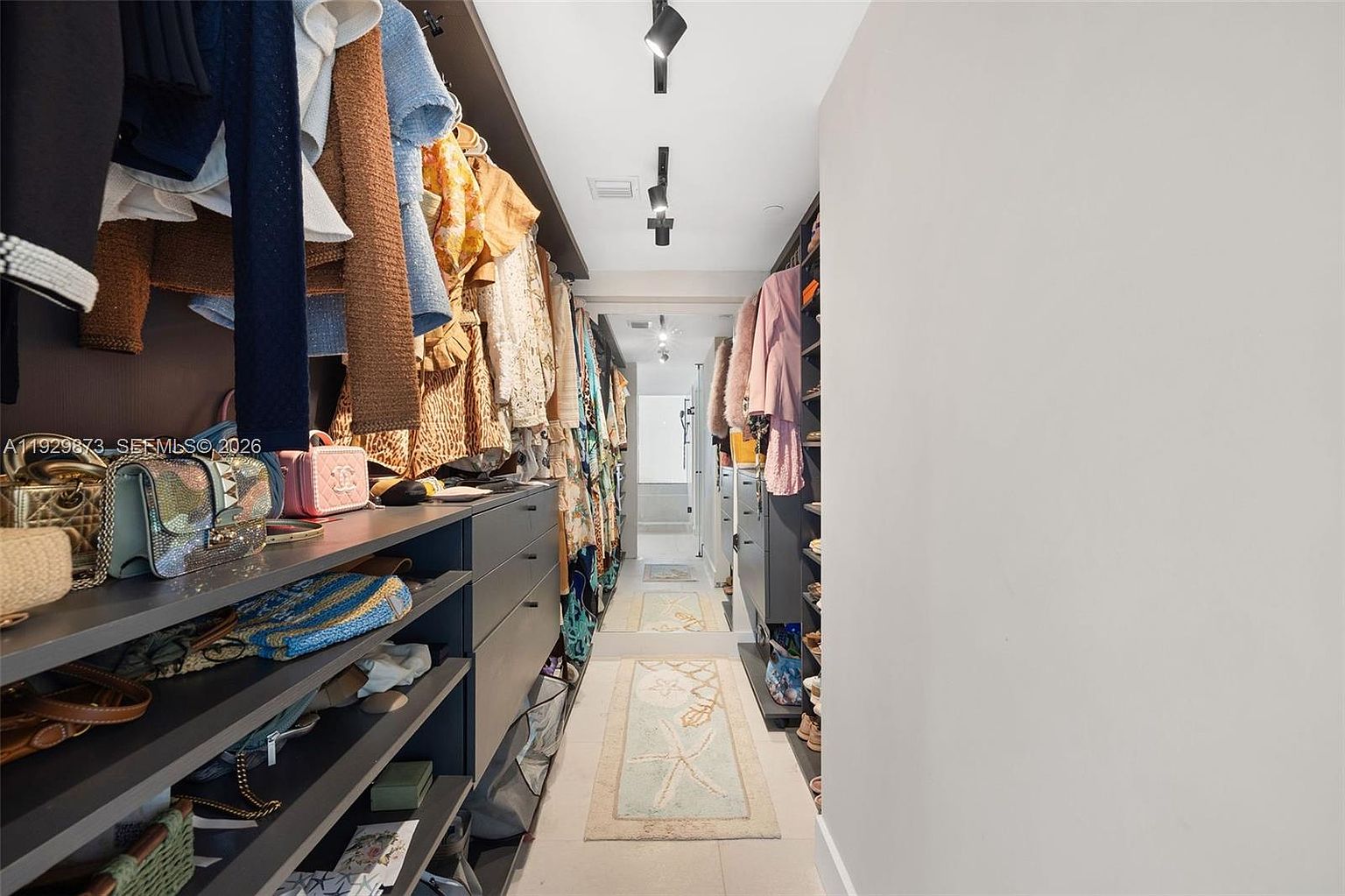 This is a well-organized walk-in closet featuring custom shelving and hanging rods filled with clothing and accessories. The closet is illuminated by track lighting, and a runner rug adds a touch of style to the tiled floor. The overall impression is one of luxury and efficient storage.