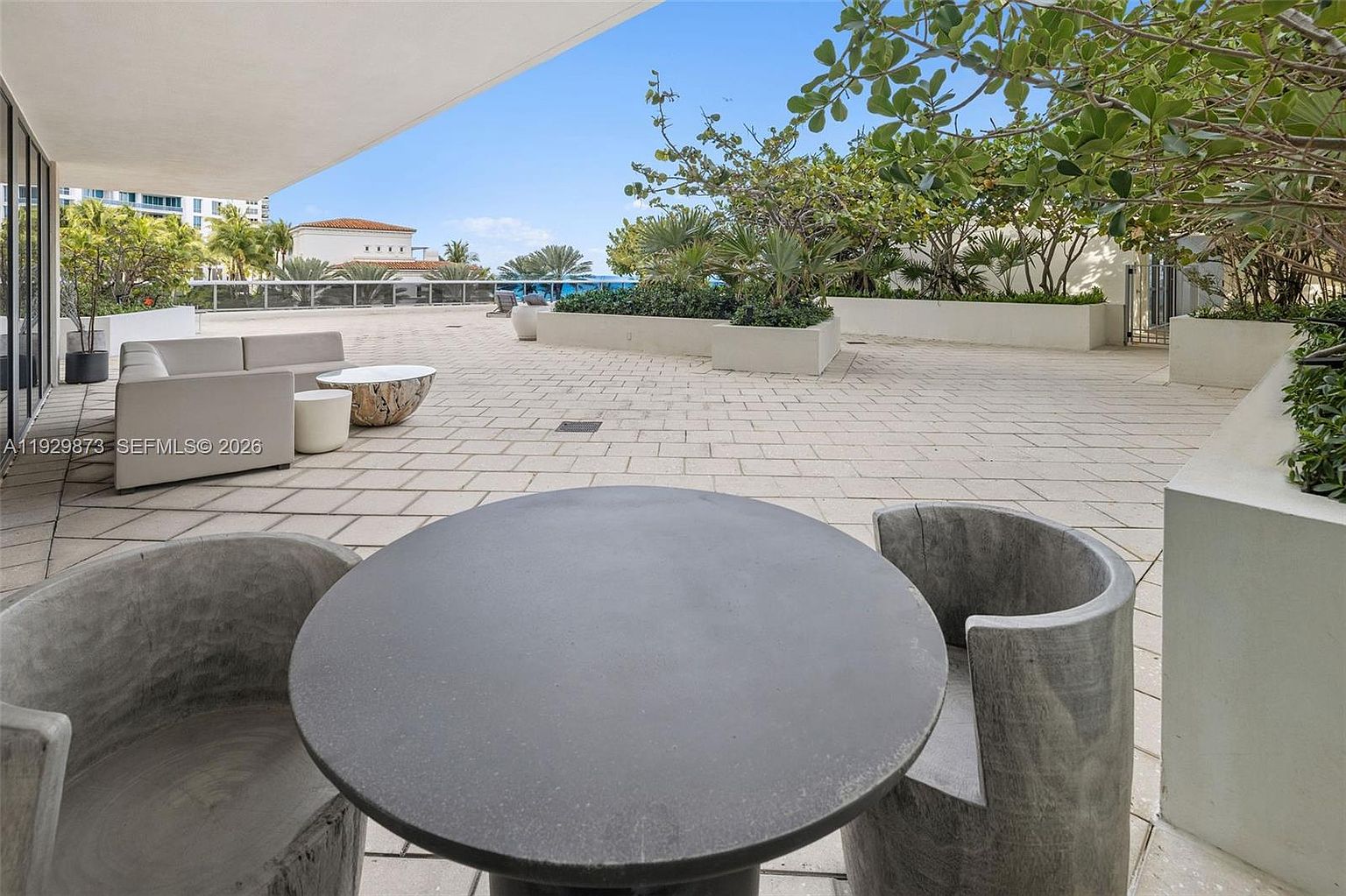 This image showcases a spacious outdoor patio or balcony area, featuring modern furniture including a round table with two unique chairs and a sectional sofa. The area is paved with light-colored stone tiles and surrounded by lush greenery in raised planters, offering a serene and private outdoor living space. The view extends to other buildings and a clear blue sky, suggesting a desirable location.