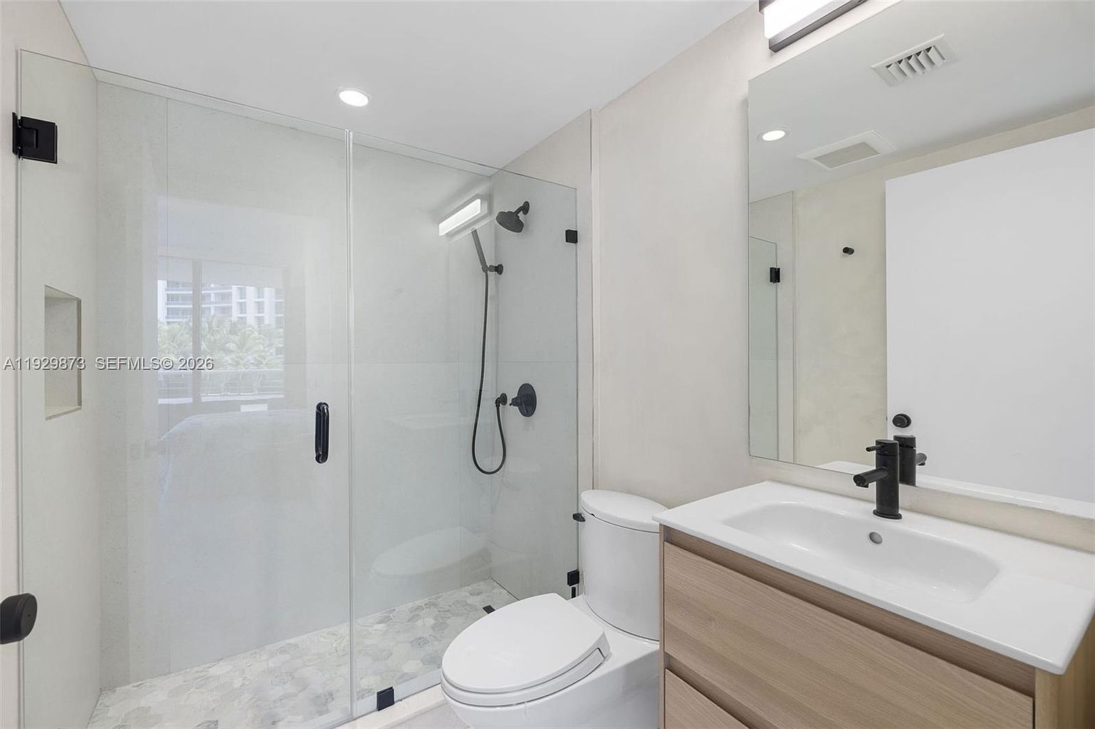 This is a modern bathroom featuring a glass-enclosed shower with black hardware, a white toilet, and a vanity with a light wood cabinet and white countertop. The walls are a neutral color, and the floor is tiled with a hexagonal pattern. The perspective is from the doorway, showcasing the entire bathroom layout.