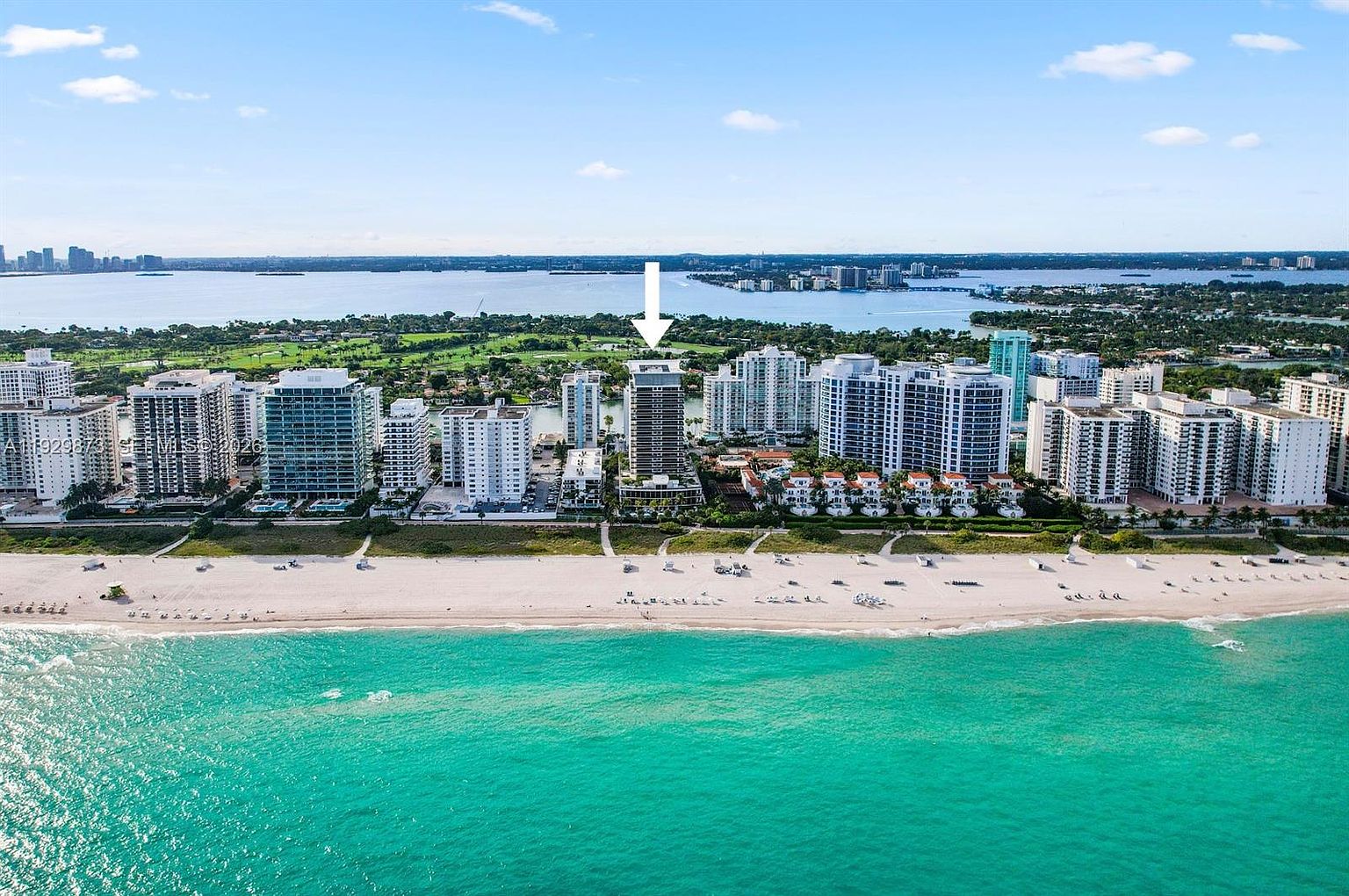 This aerial shot showcases a luxury beachfront property with a modern high-rise condominium as the focal point. The building is surrounded by lush landscaping, a pristine sandy beach, and turquoise ocean waters. The image conveys a sense of exclusivity and high-end coastal living.