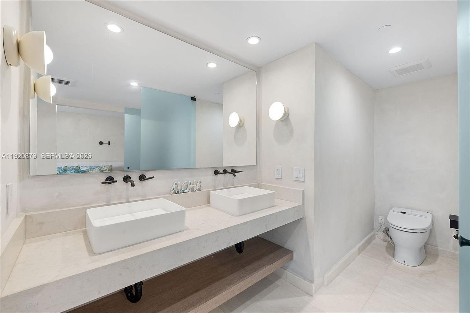 This is a bright and modern primary bathroom featuring a double vanity with white vessel sinks and matte black fixtures. A large mirror spans the length of the vanity, reflecting the clean lines and minimalist design. The toilet is a modern, elongated model, and the overall color palette is white and neutral, creating a spa-like atmosphere.