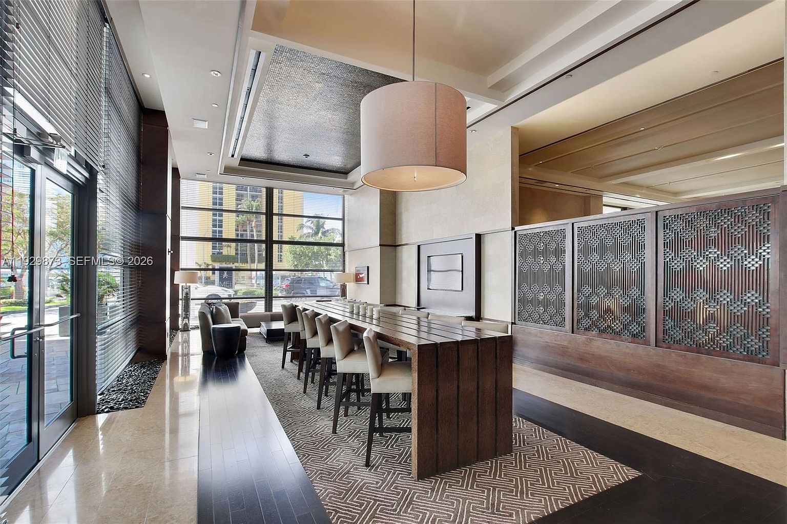 This interior shot showcases a modern and stylish living room area, possibly in a condominium or apartment building. The space features a long, wooden communal table with bar stools, a seating area with contemporary furniture, and large windows that provide ample natural light. The design incorporates a mix of textures and materials, creating a sophisticated and inviting atmosphere.
