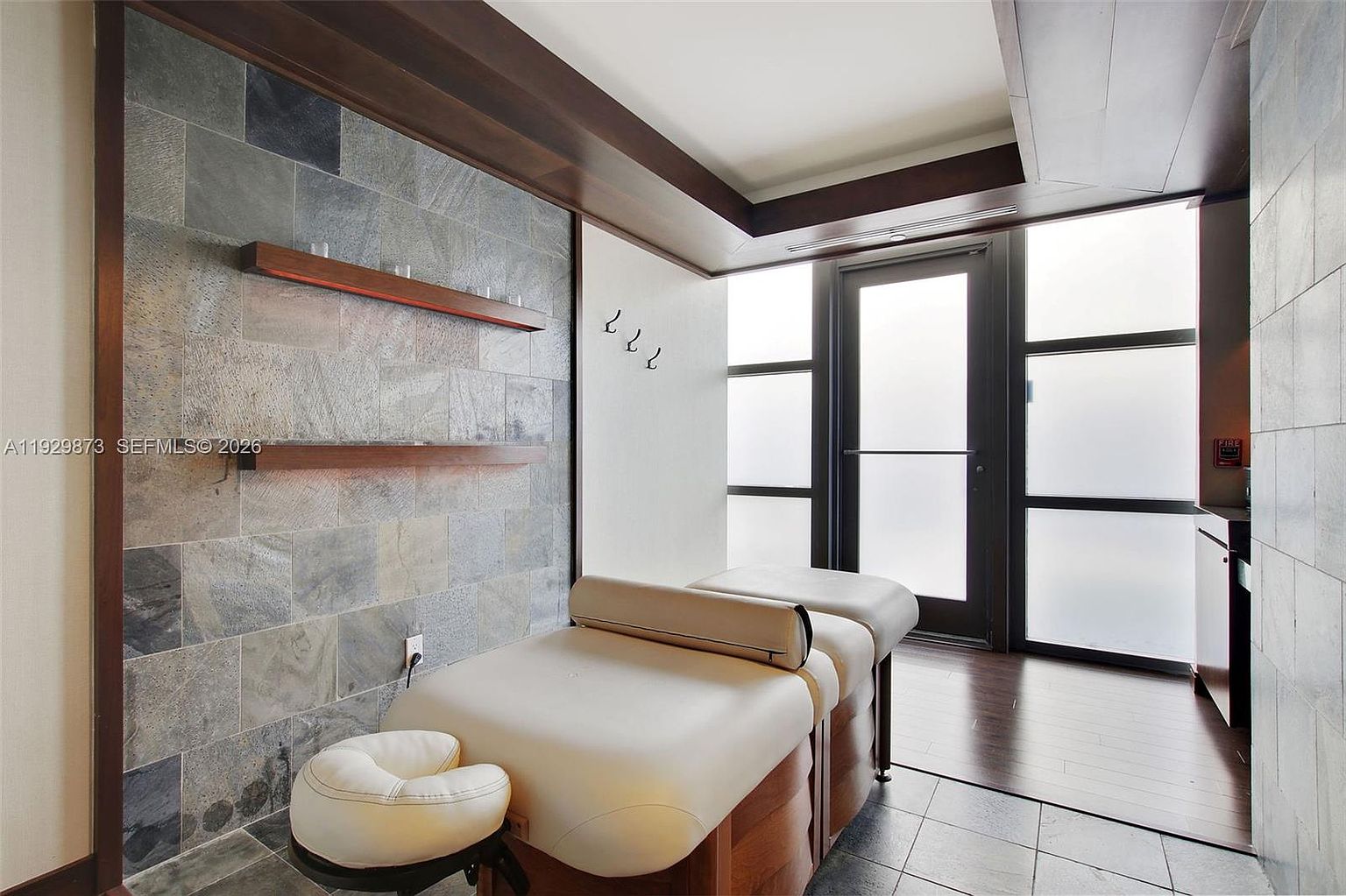 This interior shot showcases a spa or massage room, featuring a massage table with a headrest and a stool. The room has a modern aesthetic with a stone-tiled wall, wooden shelves, and a dark-framed glass door. The lighting is soft, creating a relaxing and serene atmosphere.