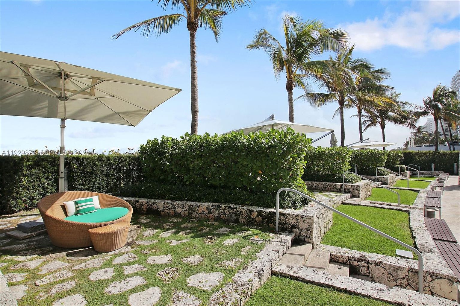 This image showcases a beautifully landscaped yard or garden area, possibly part of a larger property's amenities. The space features a stone pathway interspersed with grass, leading to tiered garden beds with lush greenery and palm trees in the background. A comfortable outdoor seating area with an umbrella provides a relaxing spot, enhancing the appeal of this outdoor space.