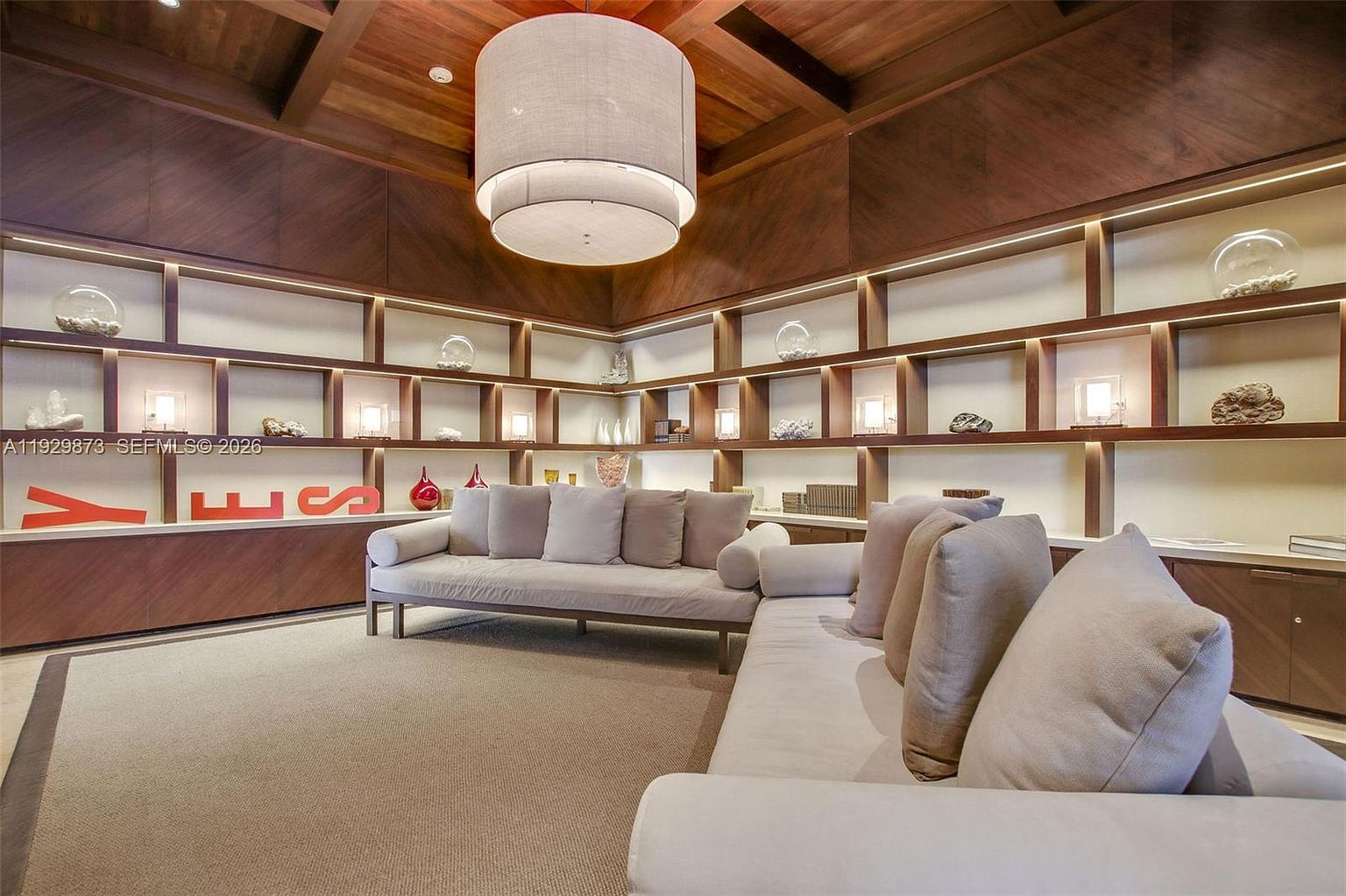 This interior shot showcases a stylish living room with a focus on modern design and comfort. The room features a large, neutral-toned sectional sofa, complemented by a custom-built shelving unit displaying decorative items and ambient lighting. A statement pendant light fixture hangs from the wooden ceiling, adding to the room's sophisticated ambiance.