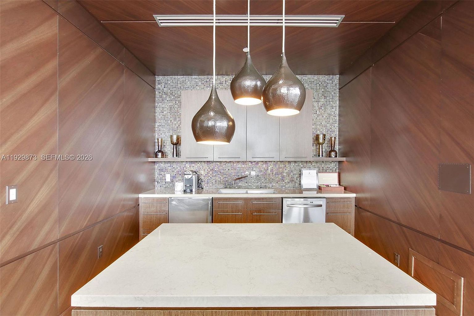 This interior shot showcases a modern kitchen area with rich wood paneling and a mosaic tile backsplash. Three teardrop-shaped pendant lights hang above a large, light-colored island, providing ample illumination. Stainless steel appliances and sleek cabinetry complete the contemporary design, creating a sophisticated and functional space.