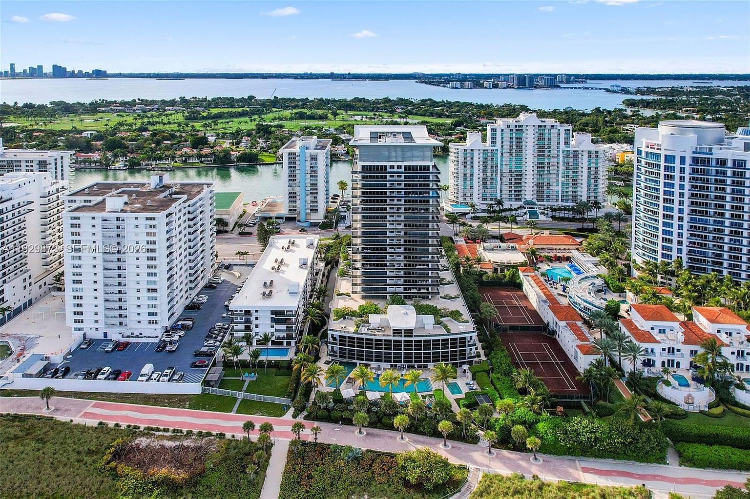 This aerial shot showcases a luxurious waterfront property featuring a modern high-rise condominium with multiple pools, tennis courts, and meticulously landscaped grounds. The building's architecture is sleek and contemporary, complemented by surrounding lush greenery and stunning water views, creating an impression of upscale coastal living.