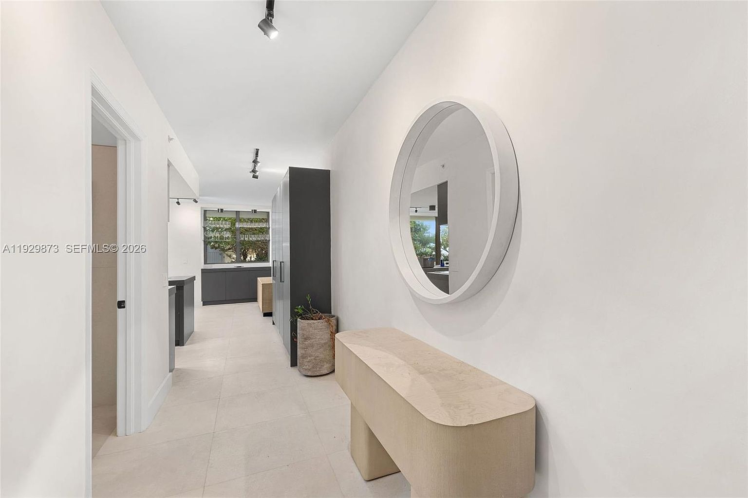 This interior shot showcases a bright and modern hallway. The space features light-colored tile flooring, white walls, and a round mirror above a stylish bench. A glimpse of the kitchen area is visible in the background, adding depth to the scene.