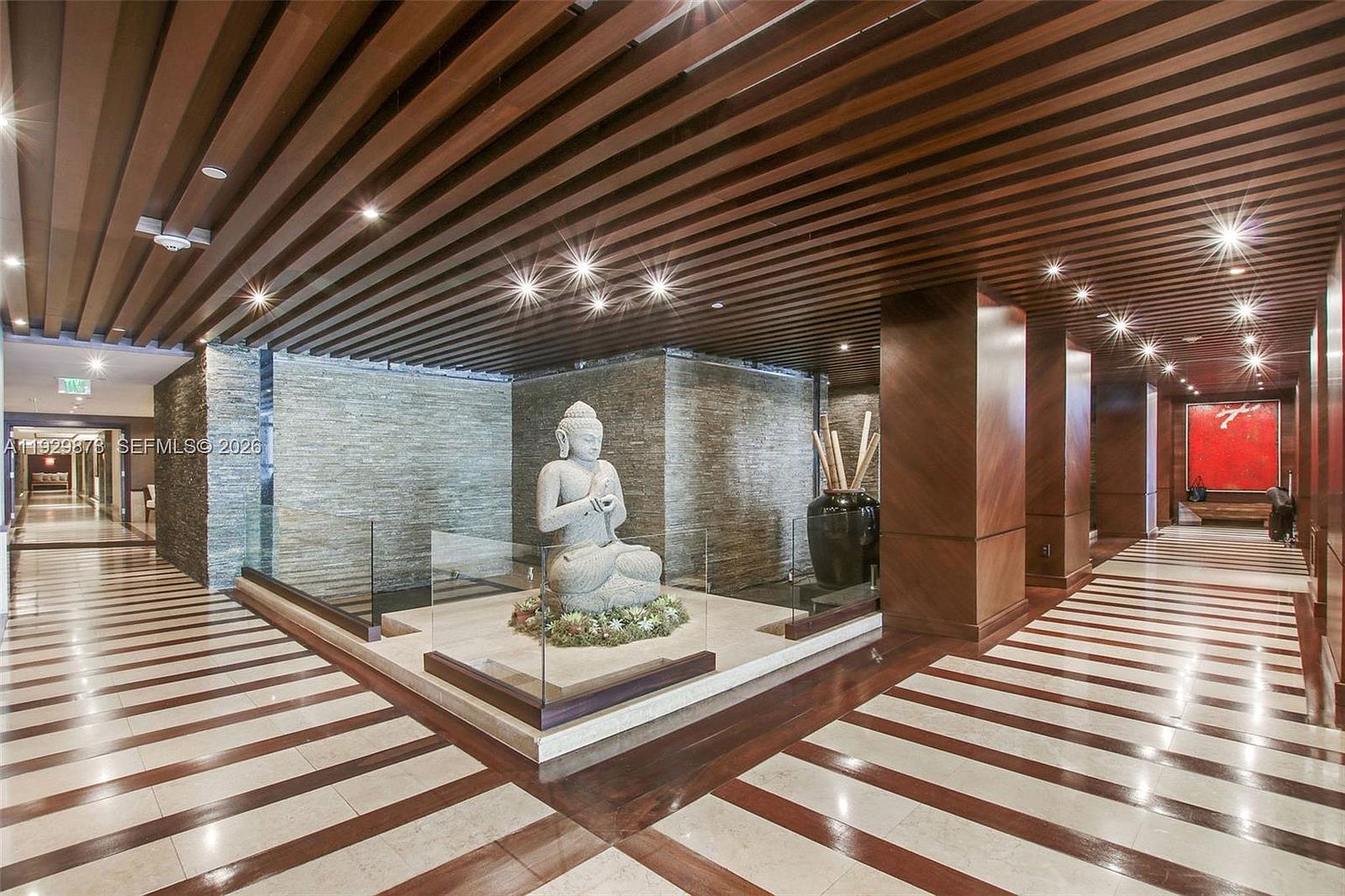 This interior shot showcases a luxurious hallway featuring a striking combination of wood and stone elements. A central Buddha statue is surrounded by glass, creating a focal point. The ceiling is adorned with wooden slats and recessed lighting, while the floor boasts a geometric pattern of light and dark wood, leading to a hallway with a red painting at the end.