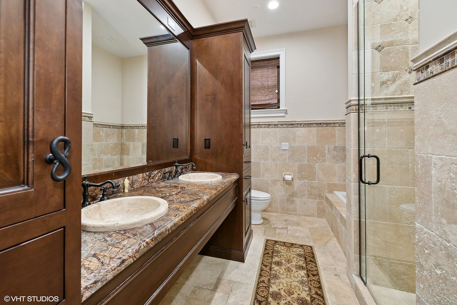 This is a primary bathroom featuring a double vanity with a granite countertop and two sinks. The cabinetry is dark wood, and the walls are tiled in a neutral stone. A glass-enclosed shower is visible on the right, and a window provides natural light. The overall impression is luxurious and well-appointed.