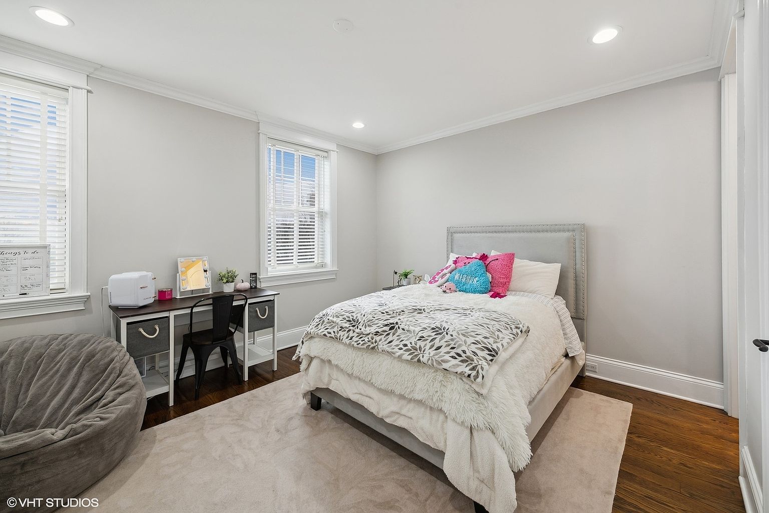 This is a cozy bedroom featuring a bed with a gray upholstered headboard, a fluffy white comforter, and decorative pillows. A desk with a black chair sits near a window, and a gray beanbag chair adds a casual touch. The room has hardwood floors and light gray walls, creating a serene and inviting atmosphere.