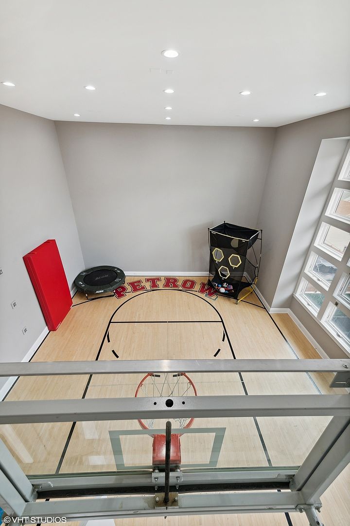 This is an interior shot of a home gym featuring a basketball half-court. The court has a wooden floor with black lines and a basketball hoop. There is also a red pad, a small trampoline, a ball rack, and the word "RETRO" painted on the court. The perspective is from above, looking down at the court.