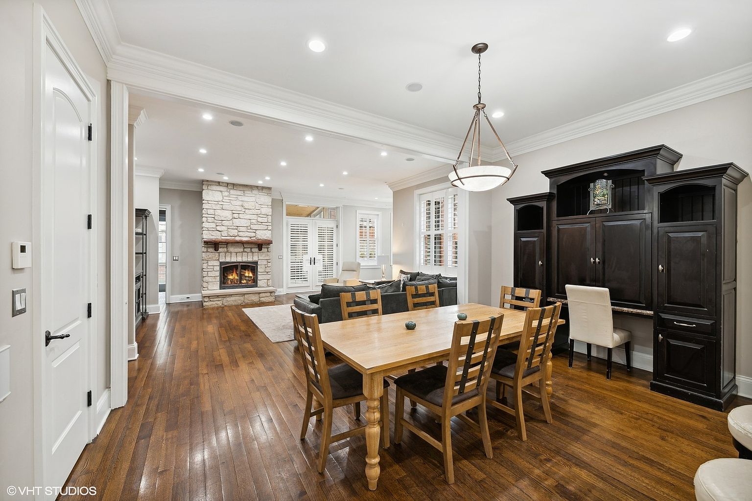 This interior shot showcases a dining room with hardwood floors and a light wood dining table set for six. A dark wood built-in cabinet stands against the wall, providing storage and a workspace. The room flows into a living area with a stone fireplace, creating an open and inviting atmosphere.